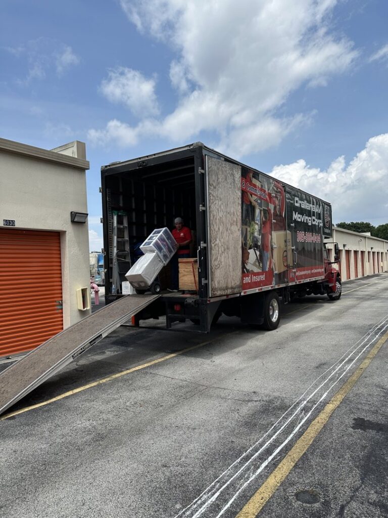 A mover from Orellana AV Moving in Miami, FL, loading items into a moving truck with an extended ramp, next to storage units.