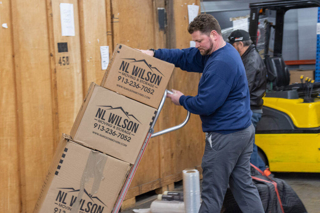 A mover from N. L. Wilson Moving and Storage pushing a hand truck with stacked boxes in a warehouse in Olathe, KS.
