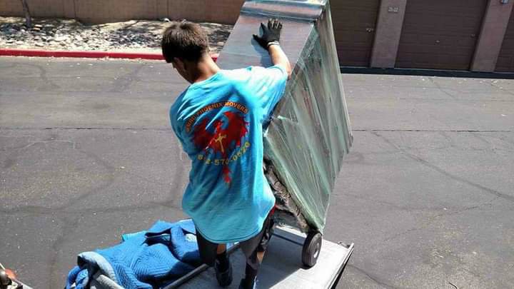 A mover from Rising Phoenix Movers LLC pushing a large, wrapped item on a dolly up a truck ramp in Phoenix, AZ.
