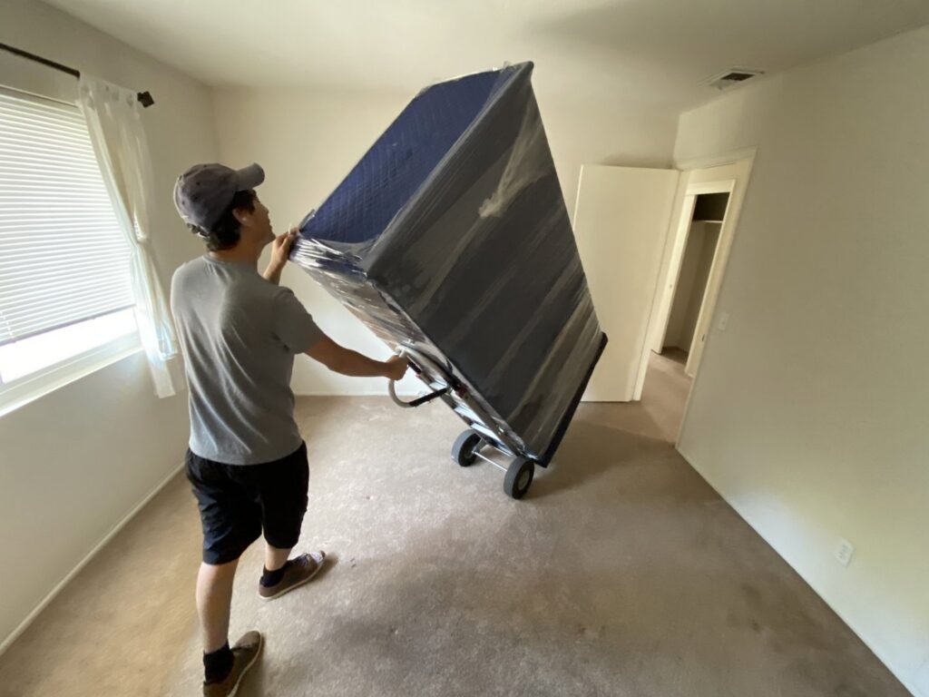 A mover from Sunshine Moving Services LLC transporting a large, wrapped piece of furniture on a dolly in San Diego, CA.
