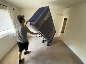 A mover from Sunshine Moving Services LLC transporting a large, wrapped piece of furniture on a dolly in San Diego, CA.