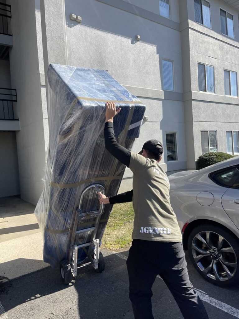 A mover from J.Gentle Van Lines transports a large, wrapped piece of furniture on a dolly outside an apartment building in Elizabeth, NJ.