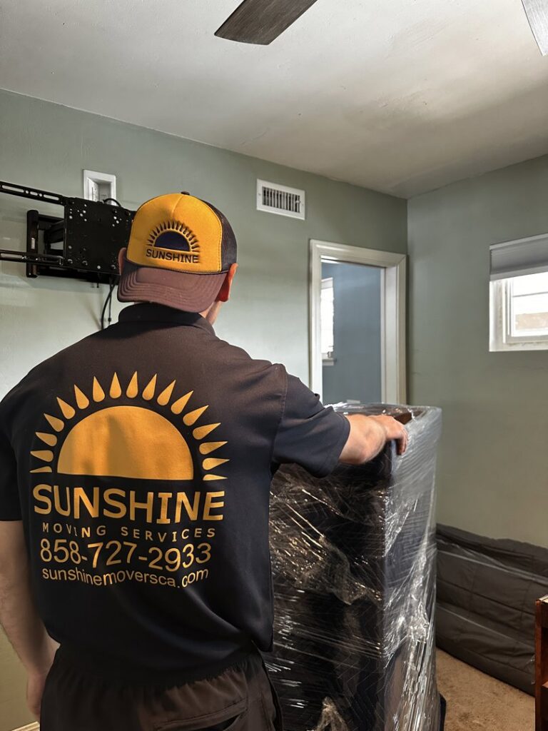 A mover from Sunshine Moving Services LLC carefully transporting a large, wrapped piece of furniture in San Diego, CA.