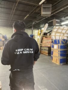 A J.Gentle Van Lines mover stands in a warehouse or storage facility surrounded by stacked items in Elizabeth, NJ.