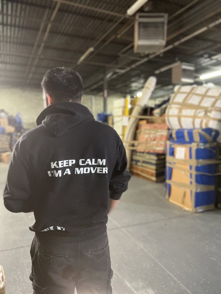 A J.Gentle Van Lines mover stands in a warehouse or storage facility surrounded by stacked items in Elizabeth, NJ.