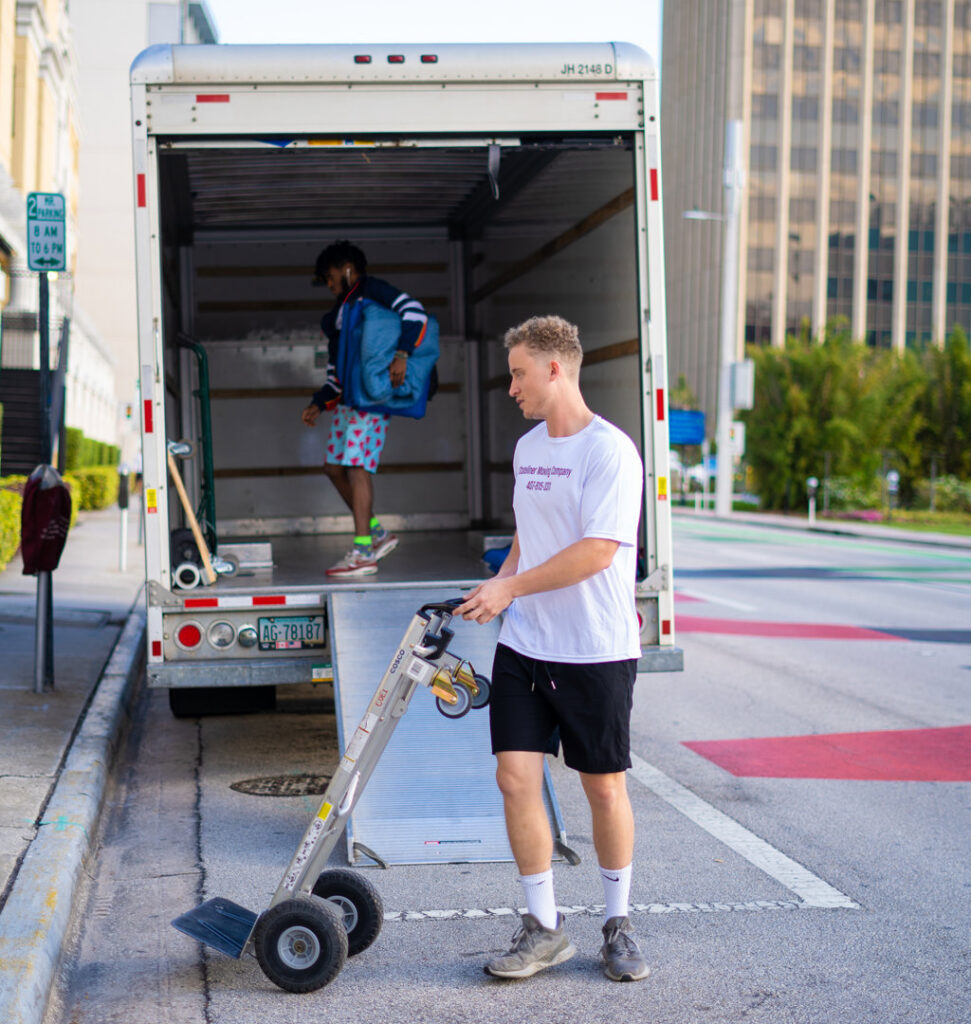 A mover from The Local Movers using a hand truck to unload items from a moving truck on a city street in Orlando, FL.