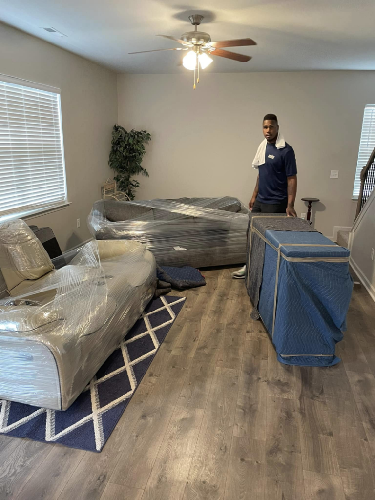 A professional mover from CNZ Moving in Raleigh, NC, standing with wrapped furniture and a dolly in a client's living room.