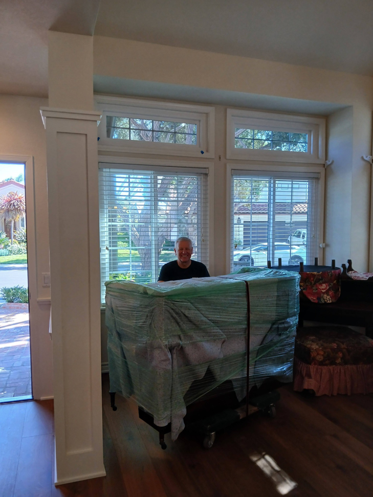 A mover standing behind a large, plastic-wrapped piece of furniture on a dolly inside a home by Rough Çutt Movers in San Diego, CA.