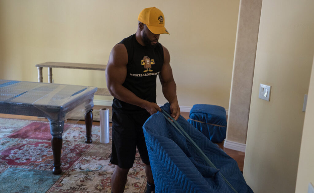 A Muscular Moving Men & Storage mover carefully wrapping furniture with a blue moving blanket inside a home in Phoenix, AZ.