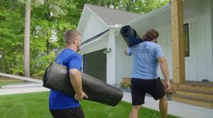 Two movers carrying rolled-up floor protectors towards a house for Goal Line Moving in Maple Grove, MN.