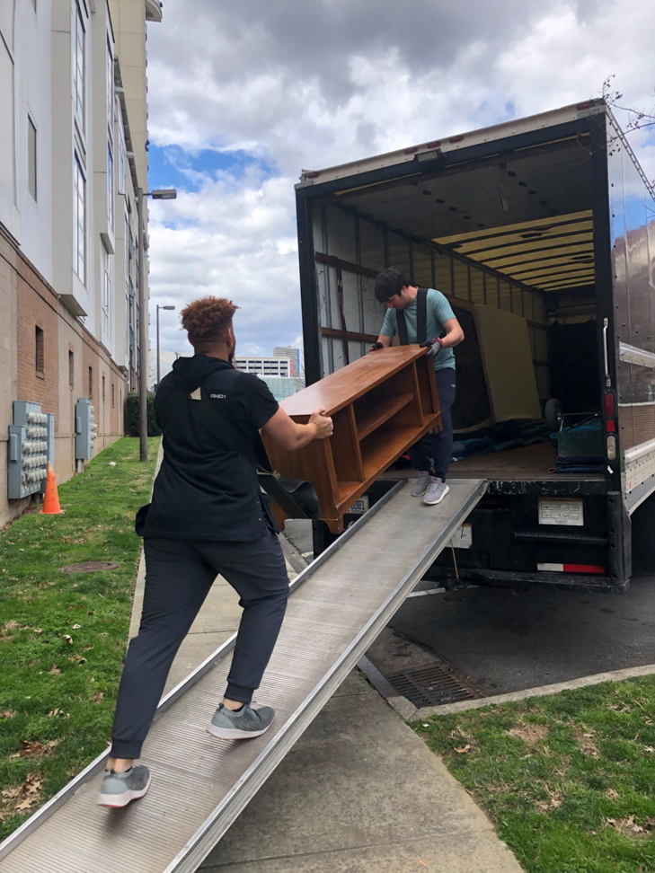 Two Roma Moving professionals carefully carrying a wooden cabinet up a ramp into a moving truck in Charlotte, NC.