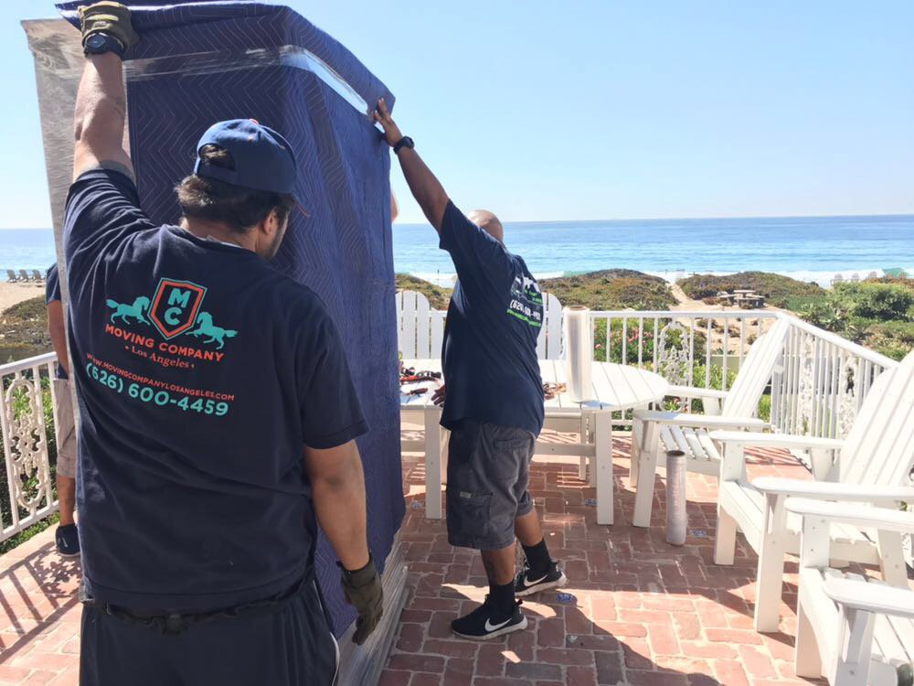 Two movers from Cheap Movers Los Angeles carrying blanket-wrapped furniture on a balcony with an ocean view in Los Angeles, CA.