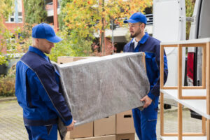 Two professional movers from Three Movers in Henderson, NV, carrying a wrapped piece of furniture towards a moving van.