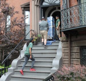 Three Mastodon Moving LLC movers carefully carrying a large, wrapped item up outdoor stairs in Westborough, MA.
