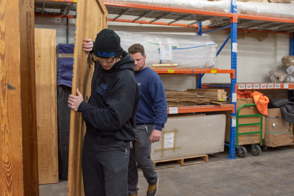 Two movers from N. L. Wilson Moving and Storage carrying a large wooden panel in a warehouse in Olathe, KS.