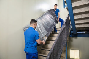 Two professional movers from Three Movers in Henderson, NV, carefully carrying a large, wrapped piece of furniture up a staircase.