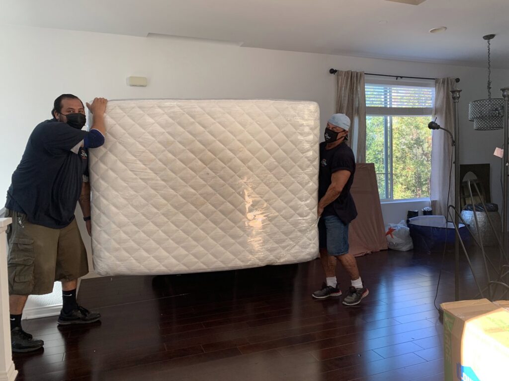 Two movers from Cheap Movers Los Angeles carefully carrying a large, plastic-wrapped mattress inside a house in Los Angeles, CA.