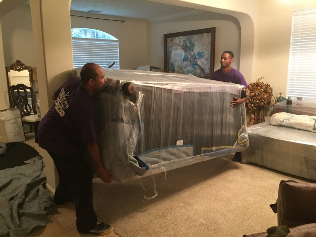 Two skilled movers from Kingdom Men Movers in Houston, TX, carefully carrying a large, wrapped sofa through a living room.
