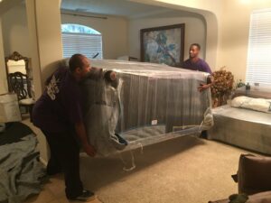 Two skilled movers from Kingdom Men Movers in Houston, TX, carefully carrying a large, wrapped sofa through a living room.