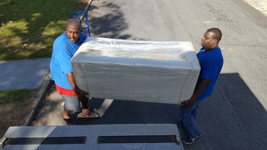Two Ron's Moving team members carrying a wrapped sofa onto a moving truck ramp in Little Rock, AR.