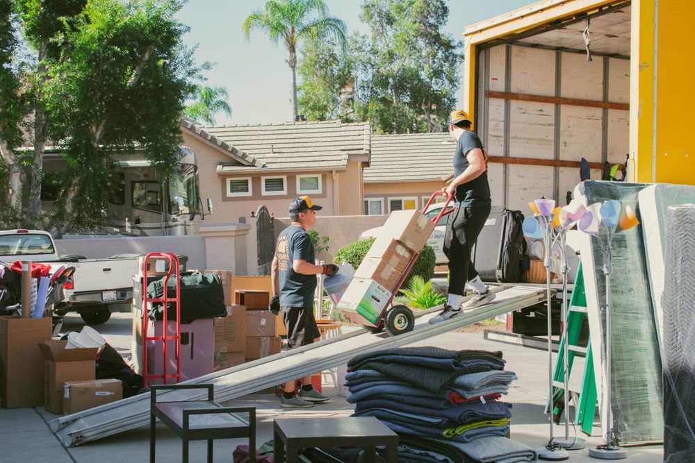 Two movers from Sunshine Moving Services LLC efficiently loading moving boxes onto a truck using a ramp in San Diego, CA.