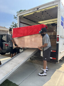 Two movers from Gifted Journey Movers loading a large, wrapped furniture item onto a moving truck via a ramp in Gulfport, MS.