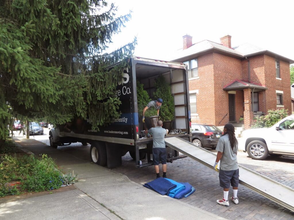 Three movers from Leaders Moving & Storage Co. loading potted plants onto a moving truck via a ramp in Indianapolis, IN.