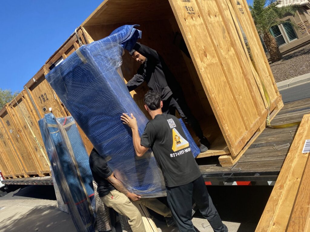 Muscular Moving Men & Storage movers carefully loading a wrapped sofa into a large wooden crate on a truck in Phoenix, AZ.