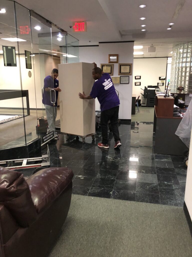 Two movers from Kingdom Men Movers in Houston, TX, carefully moving a large, wrapped item on a dolly through an office space.
