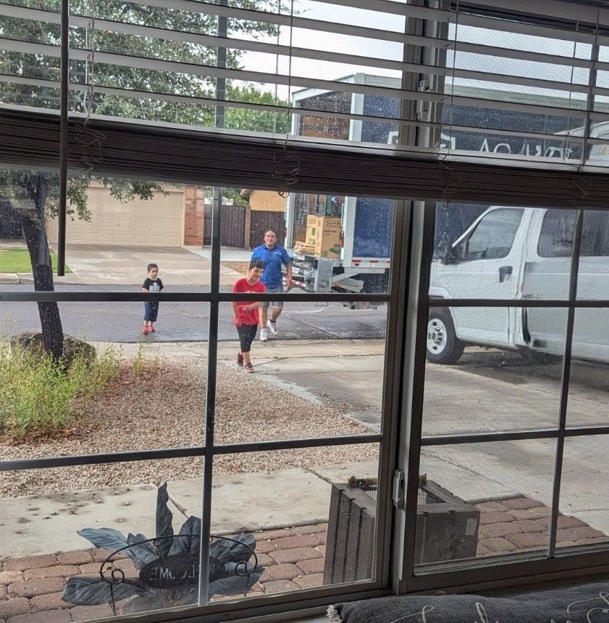 A view through a window showing Academy Movers personnel and a moving truck outside a residence in Gilbert, AZ.