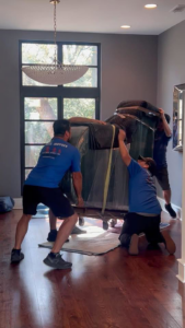 Three professional movers from Antz Movers Company in Austin, TX, carefully relocating a large, wrapped piece of furniture inside a home.