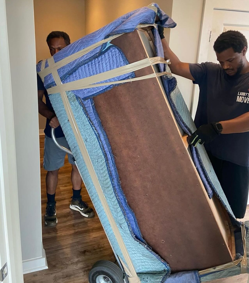 Two movers from Larry's Crew Movers in Atlanta, GA, transporting a large, wrapped piece of furniture on a dolly.