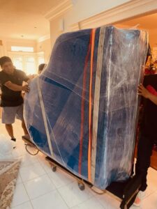 Professional movers transporting a large, wrapped piano on a dolly inside a home for Emanuel Movers, Inc. in Houston, TX