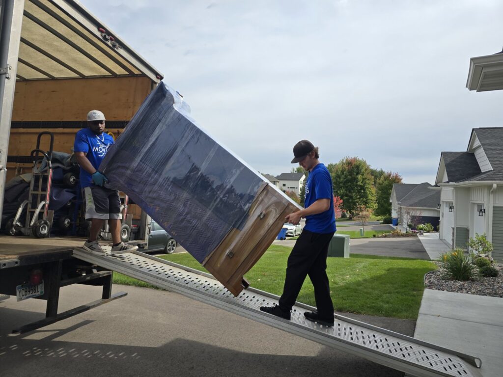 Two movers unloading a large, wrapped dresser down a ramp from a moving truck for Goal Line Moving in Maple Grove, MN.