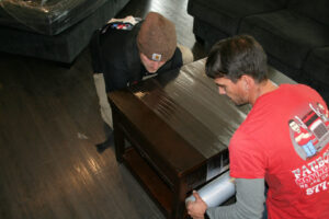 Two movers wrapping a coffee table with protective plastic for Fatboy Slims Moving Company in Colorado Springs, CO.