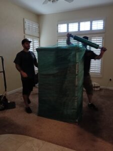 Two movers from Rising Phoenix Movers LLC carefully wrapping a large piece of furniture inside a home in Phoenix, AZ.