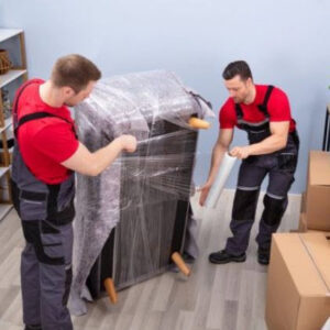 Two professional movers from Three Movers in Henderson, NV, carefully wrapping a piece of furniture with plastic for protection.