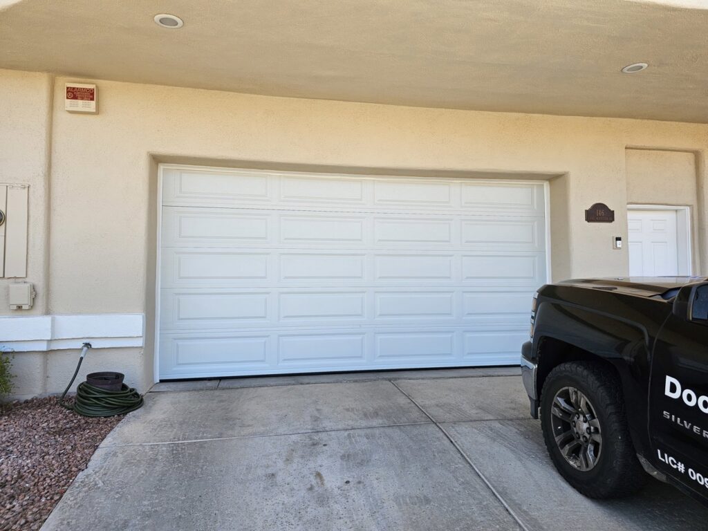 A newly installed white panel garage door on a house, with a work truck nearby, by Rebel Overhead Doors in Las Vegas, NV.