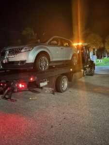 A black flatbed tow truck from Always Ready Towing transporting a silver Lincoln SUV at night in North Fort Myers, FL.
