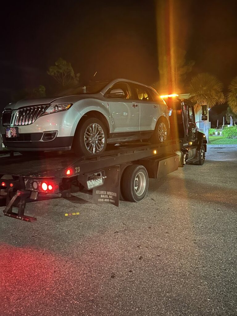 A black flatbed tow truck from Always Ready Towing transporting a silver Lincoln SUV at night in North Fort Myers, FL.