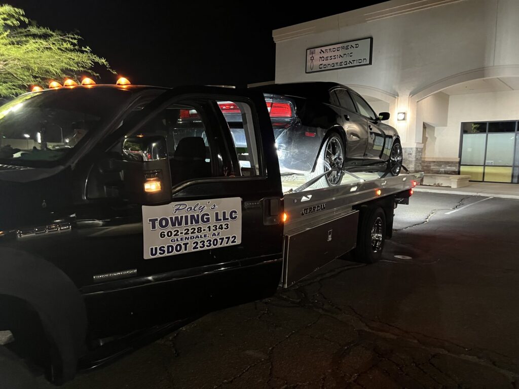 A black flatbed tow truck from Poly's Towing Glendale, AZ, providing night towing service for a black sedan.