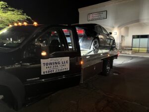 A black flatbed tow truck from Poly's Towing Glendale, AZ, providing night towing service for a black sedan.