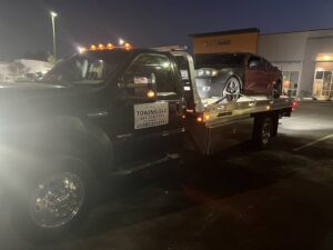 A black flatbed tow truck from Poly's Towing Glendale, AZ, providing night towing service for a dark sedan.