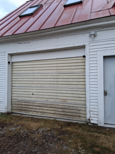 An old and dirty roll-up garage door on a white building, ready for service from Ski Door, Inc. in Center Rutland, VT.