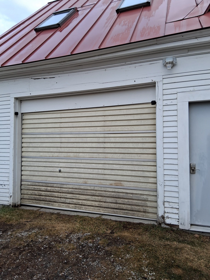 An old and dirty roll-up garage door on a white building, ready for service from Ski Door, Inc. in Center Rutland, VT.