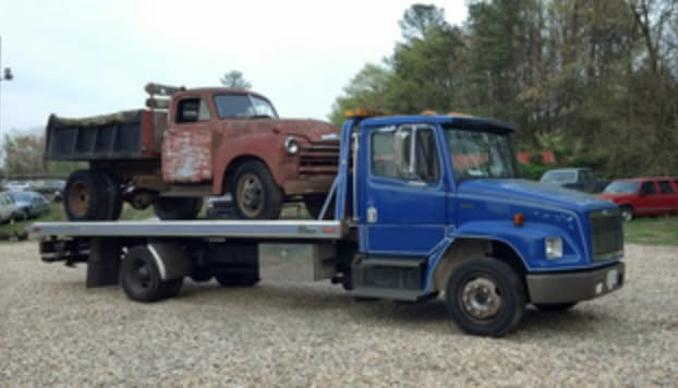An old, rusty dump truck being transported on a blue flatbed tow truck by Bakersfield Towing Company in Bakersfield, CA.