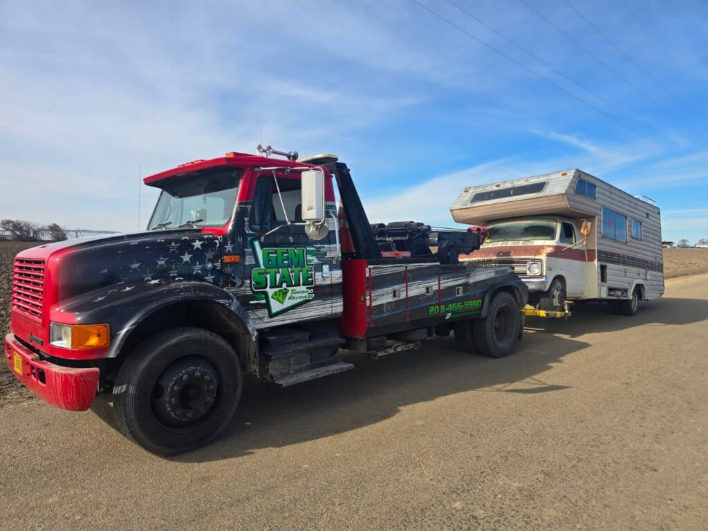 An older RV camper being towed by a wrecker from Gem State Towing and Recovery in Caldwell, ID.
