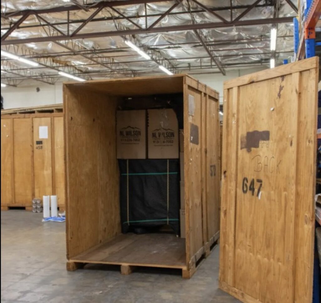 An open wooden storage crate with boxes and a wrapped item inside, from N. L. Wilson Moving and Storage in Olathe, KS.