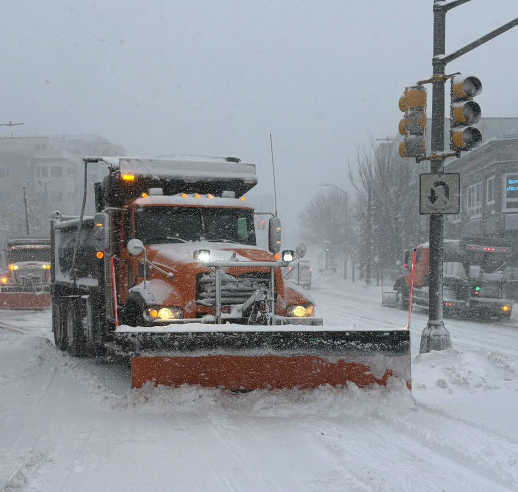 An orange snow plow truck clearing a street during a snowfall for the City of Cambridge Department of Public Works in Cambridge, MA.