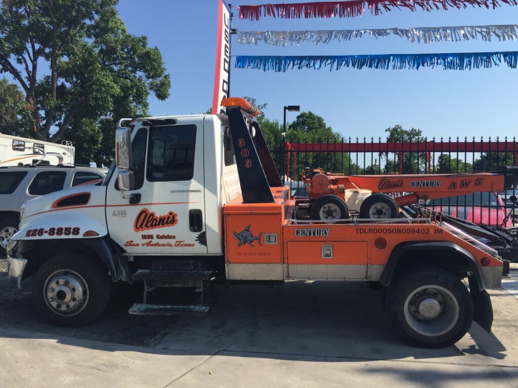 An orange and white wrecker truck with Alanis Wrecker Service branding parked in San Antonio, TX.